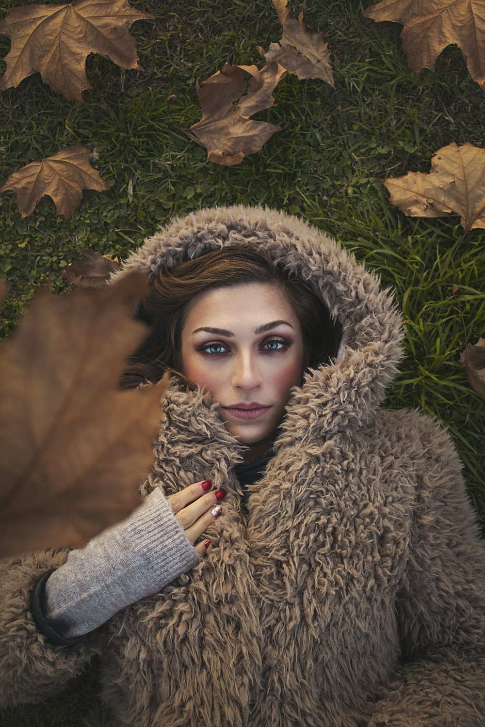 woman wearing brown fur zip-up jacket lying on green field surrounded with brown leaves