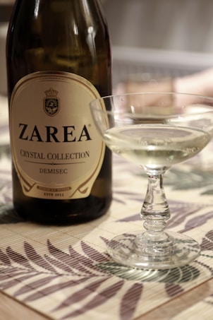 A bottle of Zarea Crystal Collection Demisec wine is placed on a patterned surface next to a filled wine glass. The bottle has a crest and detailed label, and the wine in the glass has a light, clear color.