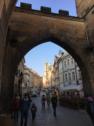A historic city street viewed through a stone archway, with people walking along a cobblestone path lined by old buildings. The architecture features ornate facades and a church or cathedral with twin towers in the background. The street is lively with various pedestrians and some outdoor seating areas visible.