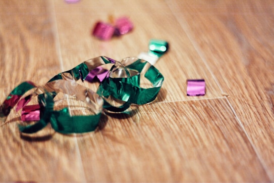 Colorful confetti and streamers are scattered on a wooden floor, creating a festive and casual atmosphere. The shiny materials reflect light, and their vibrant colors contrast with the warm tones of the wood.