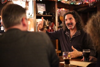 A group of people are gathered around a bar table, engaging in animated conversation. The man facing the camera has curly hair and is gesturing with his hand, possibly sharing a story. Glasses of dark beverages are on the table, and there is a warm, cozy atmosphere with soft lighting and festive decorations.
