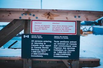 A warning sign posted on a wooden structure instructs individuals entering or returning to Canada to declare all weapons to a Canada Border Services Agency officer. The sign is prominently written in both English and French, with distinctive white and red lettering on a black background.