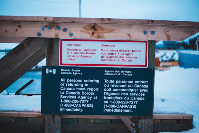 A warning sign posted on a wooden structure instructs individuals entering or returning to Canada to declare all weapons to a Canada Border Services Agency officer. The sign is prominently written in both English and French, with distinctive white and red lettering on a black background.