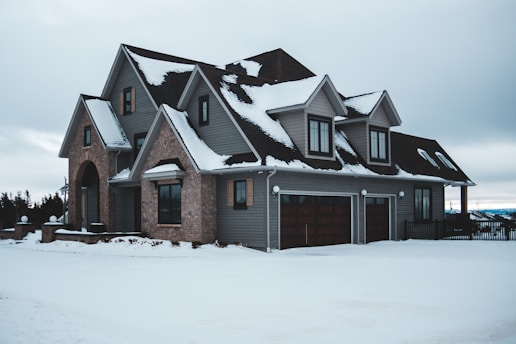 A snow-covered, large two-story house with dark brown and gray exterior, including multiple gabled windows, a garage, and a stone archway entrance. Overcast skies and a landscape with some trees are visible in the background.