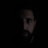 A person with a beard is partially visible, shrouded in darkness with only part of their face illuminated. The image has a mysterious and moody atmosphere, highlighting one eye and the contours of their face.