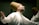 A person is performing a spiritual dance, wearing a traditional white robe and a tall brown hat. The expression on the person's face is serene and blissful, with arms outstretched in an embracing motion.