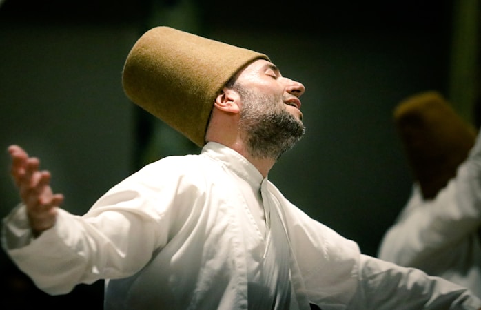 A person is performing a spiritual dance, wearing a traditional white robe and a tall brown hat. The expression on the person's face is serene and blissful, with arms outstretched in an embracing motion.