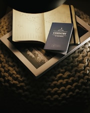 A cozy scene features a soft, woven surface upon which lies an open notebook displaying a world map with marked locations. A blue travel journal titled 'Another Country in the Books' rests atop a closed book or magazine with a monochrome landscape photo on its cover.