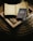 A cozy scene features a soft, woven surface upon which lies an open notebook displaying a world map with marked locations. A blue travel journal titled 'Another Country in the Books' rests atop a closed book or magazine with a monochrome landscape photo on its cover.