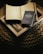 A cozy scene features a soft, woven surface upon which lies an open notebook displaying a world map with marked locations. A blue travel journal titled 'Another Country in the Books' rests atop a closed book or magazine with a monochrome landscape photo on its cover.