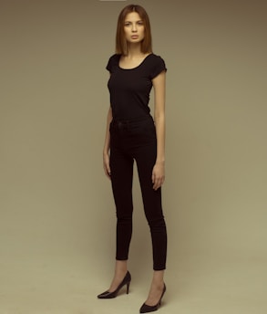 A person stands in a relaxed, confident pose, wearing a black outfit consisting of a fitted t-shirt, skinny jeans, and high heels. The background is neutral with a grayish-beige tone, creating a simple and focused composition.