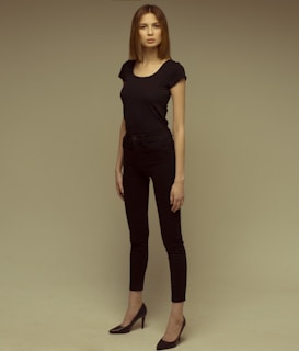 A person stands in a relaxed, confident pose, wearing a black outfit consisting of a fitted t-shirt, skinny jeans, and high heels. The background is neutral with a grayish-beige tone, creating a simple and focused composition.