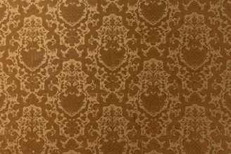 A vintage-style wallpaper with an intricate, repeating floral and damask pattern in shades of brown and beige. The design features ornate, symmetrical motifs that create a sense of elegance and classic decor.