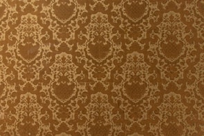 A vintage-style wallpaper with an intricate, repeating floral and damask pattern in shades of brown and beige. The design features ornate, symmetrical motifs that create a sense of elegance and classic decor.