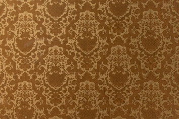 A vintage-style wallpaper with an intricate, repeating floral and damask pattern in shades of brown and beige. The design features ornate, symmetrical motifs that create a sense of elegance and classic decor.