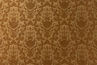 A vintage-style wallpaper with an intricate, repeating floral and damask pattern in shades of brown and beige. The design features ornate, symmetrical motifs that create a sense of elegance and classic decor.