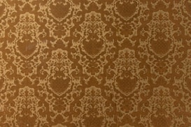 A vintage-style wallpaper with an intricate, repeating floral and damask pattern in shades of brown and beige. The design features ornate, symmetrical motifs that create a sense of elegance and classic decor.
