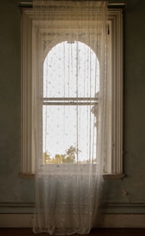 A tall, arched window is adorned with sheer, decorative curtains featuring a floral pattern. A view of trees and faint sunlight is visible through the glass, adding a serene touch to the interior space.