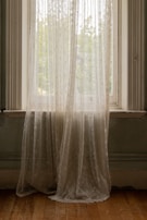 Sheer, delicate curtains hang in front of a window, gently filtering soft natural light into the room. The embroidered patterns on the fabric create a subtle, elegant texture against the backdrop of an overcast sky and faint view of greenery outside. Wooden floorboards add a touch of warmth to the subdued tones of the interior.