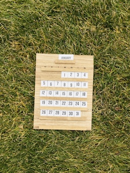 A wooden calendar displaying the month of January is placed on a lush green grass surface. The calendar is detailed with a simple, minimalist design, showing days arranged in a grid format.