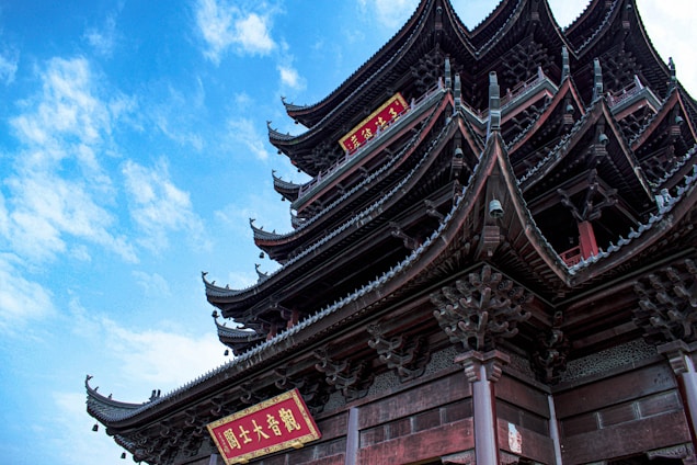 An ornate, multi-tiered pagoda with intricate architectural details, featuring curved roofs with upturned eaves. The structure displays dark wooden tones and has vibrant red signs with gold characters, set against a bright blue sky dotted with clouds.