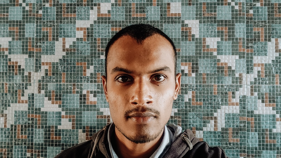 A person stares directly at the camera with a neutral expression. The background consists of a mosaic tile pattern featuring various shades of green, brown, and white.