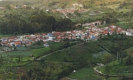 A small village with clustered houses, primarily featuring red-tiled roofs, is nestled within a lush, green landscape. Surrounding the village are terraced fields, trees, and patches of vegetation, suggesting agricultural activity. The scene is set on a hillside, providing an elevated view of the village and the surrounding natural environment.