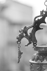 An ornate metal object with intricate designs and hanging elements. The focus is on a decorative bracket with embellishments, likely part of a larger structure. The background is blurred, enhancing the emphasis on the metalwork.