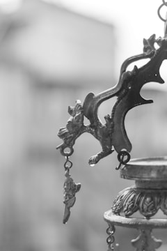 An ornate metal object with intricate designs and hanging elements. The focus is on a decorative bracket with embellishments, likely part of a larger structure. The background is blurred, enhancing the emphasis on the metalwork.