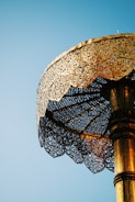 A intricately designed gold umbrella or canopy set against a clear blue sky. The structure features ornate patterns and decorative elements that reflect light, creating a stunning visual effect.