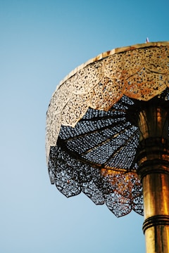 A intricately designed gold umbrella or canopy set against a clear blue sky. The structure features ornate patterns and decorative elements that reflect light, creating a stunning visual effect.