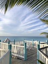 A serene beach scene featuring a wooden deck with white railings and turquoise posts, leading to a view of the ocean. Palm leaves frame the top of the image. Adirondeck chairs and a sun umbrella are positioned on the deck, enhancing the relaxing atmosphere. The sky is partly cloudy, adding to the tranquil setting.