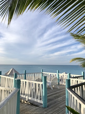 A serene beach scene featuring a wooden deck with white railings and turquoise posts, leading to a view of the ocean. Palm leaves frame the top of the image. Adirondeck chairs and a sun umbrella are positioned on the deck, enhancing the relaxing atmosphere. The sky is partly cloudy, adding to the tranquil setting.