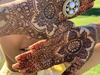 Intricate henna designs cover the hands in elaborate floral and geometric patterns. A decorative accessory with pearls and white stones is placed on one hand. The background is a blurred green, indicating a natural outdoor setting. Part of a face is visible, smiling slightly.