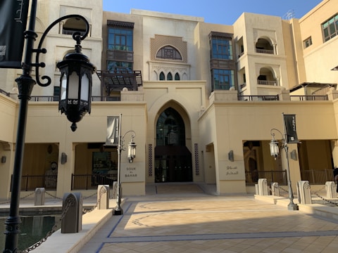 An ornate building with Middle Eastern architectural features including arched doorways and intricate window designs. The facade is cream-colored, and there are large decorative lanterns in the foreground. The entrance is marked with the words 'Souk Al Bahar' in both English and Arabic.