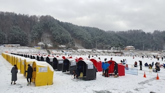 Numerous tents are set up on a snowy field surrounded by forested hills, with people engaging in outdoor activities like ice fishing. The trees are covered in snow, and the sky is overcast, creating a wintry atmosphere. Yellow and red tents are arranged in rows, and individuals are walking around, dressed in winter clothing.