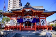 A traditional Japanese shrine with ornate architecture featuring a grey-tiled roof and red pillars. The entrance is adorned with purple banners and decorative items, and there are large brushstroke characters on a white panel. A Japanese flag flutters from a pole to the side, and the scene is set against a backdrop of modern buildings and trees.