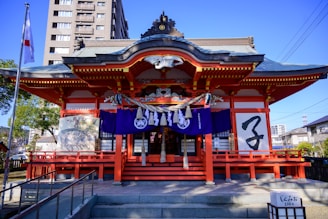 A traditional Japanese shrine with ornate architecture featuring a grey-tiled roof and red pillars. The entrance is adorned with purple banners and decorative items, and there are large brushstroke characters on a white panel. A Japanese flag flutters from a pole to the side, and the scene is set against a backdrop of modern buildings and trees.