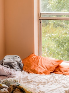 An unmade bed with white sheets and two vibrant orange pillows sits beside a window. A gray blanket is casually draped on the bed. Lush green trees are visible outside the window, providing a peaceful view and natural light.
