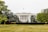 A grand, white neoclassical mansion with large columns and multiple windows stands majestically among well-maintained gardens. A flag flies atop the building, and a fountain is situated in the foreground, surrounded by lush greenery and trees.
