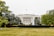 A grand, white neoclassical mansion with large columns and multiple windows stands majestically among well-maintained gardens. A flag flies atop the building, and a fountain is situated in the foreground, surrounded by lush greenery and trees.