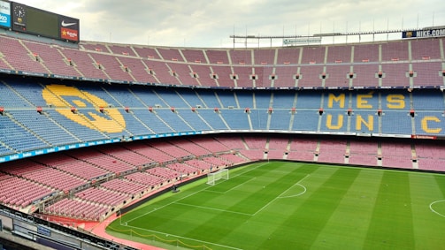 A large empty football stadium with multi-colored seating. The seats are arranged in patterns that spell out phrases. The grass pitch is well-maintained and there are advertisements around the stadium.