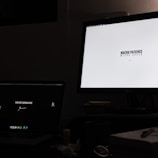 A dimly lit room with a desk containing two computer monitors. The left screen displays a message on a dark background, saying 'YOU'RE GONNA DIE', while the right monitor shows a white background with the text 'MACRO PATIENCE, MICRO SPEED'. Below the monitors, there are scattered items such as a cup, a notebook, and some papers or tissues.