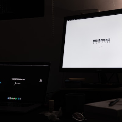 A dimly lit room with a desk containing two computer monitors. The left screen displays a message on a dark background, saying 'YOU'RE GONNA DIE', while the right monitor shows a white background with the text 'MACRO PATIENCE, MICRO SPEED'. Below the monitors, there are scattered items such as a cup, a notebook, and some papers or tissues.