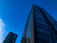 A modern skyscraper with reflective glass windows stretches upwards against a clear blue sky. The building's surface reflects the sky, creating an illusion of transparency. Adjacent, another high-rise is partially visible, contributing to a sense of urban development and scale.