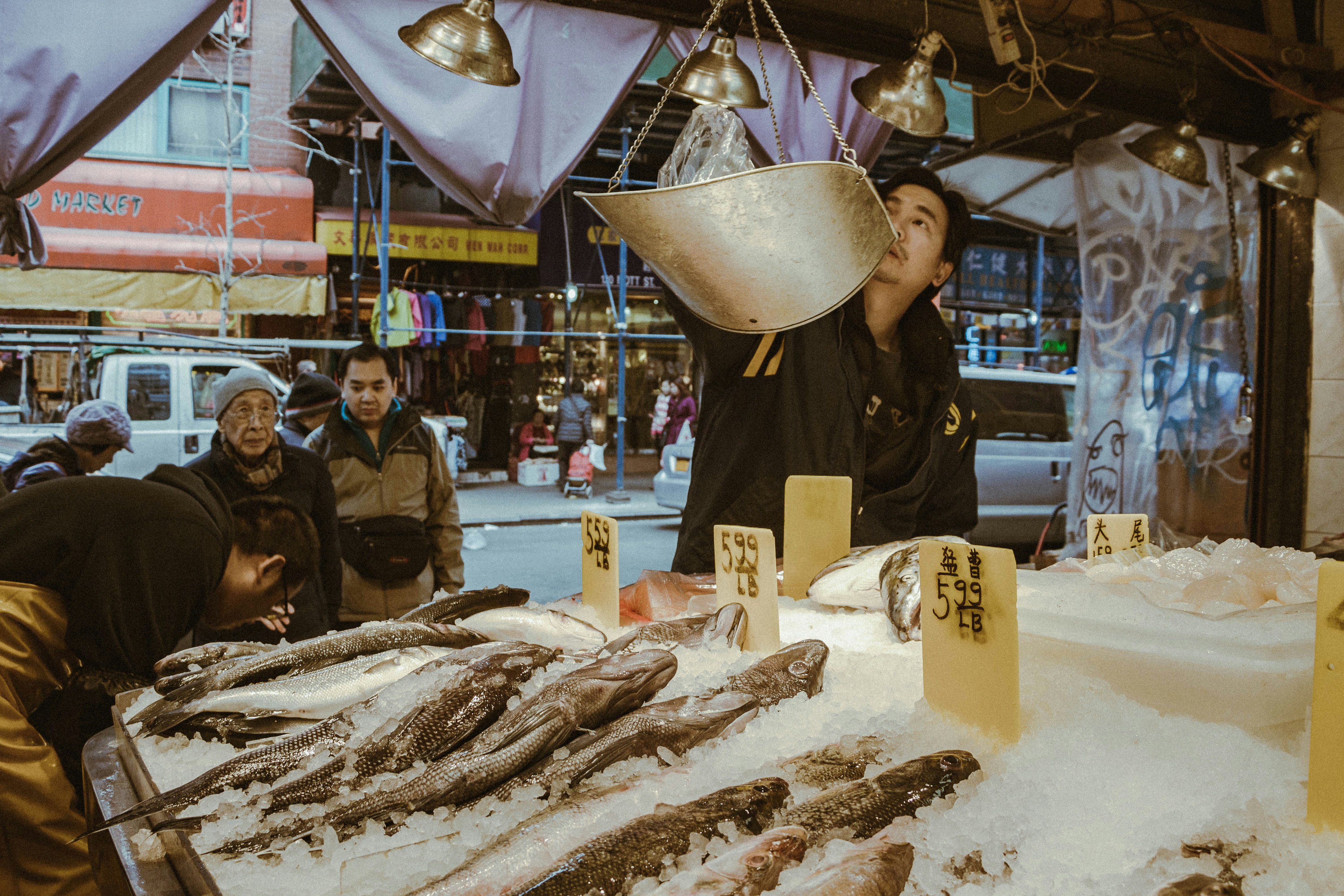 Photo of man weighing fish in Chinatown New York Manhattan with Purple sheets hanging from the ceiling. Different types of fish and there prices sit on ice in forground.