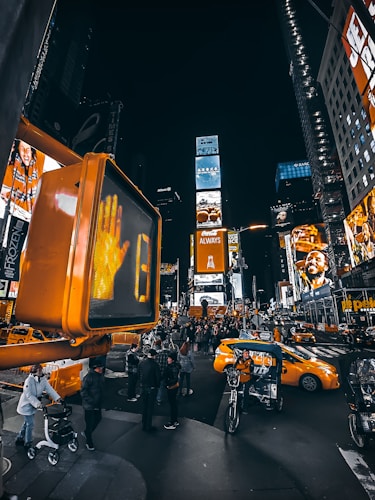 A busy urban scene at night with bright digital billboards and advertisements lighting up the area. A large pedestrian signal, showing the 'don't walk' hand, prominently stands in the foreground. Several people are gathered on the street, some walking or on bicycles. Yellow taxis and other vehicles navigate the bustling intersection.