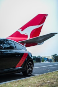 A black car with red stripes is parked near an airplane. The airplane's tail with a distinctive red and white emblem is prominent in the background, indicating proximity to an airport area.