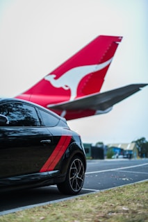 A black car with red stripes is parked near an airplane. The airplane's tail with a distinctive red and white emblem is prominent in the background, indicating proximity to an airport area.