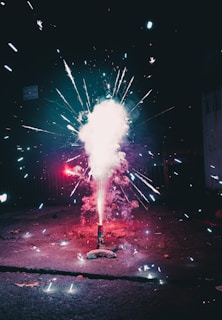 A vibrant fireworks display with sparks and colorful lights emanating from a singular source on the ground. The area is dimly lit, highlighting the contrast between the bright fireworks and the dark surroundings. Vibrant colors in the explosion create a dynamic and captivating scene.
